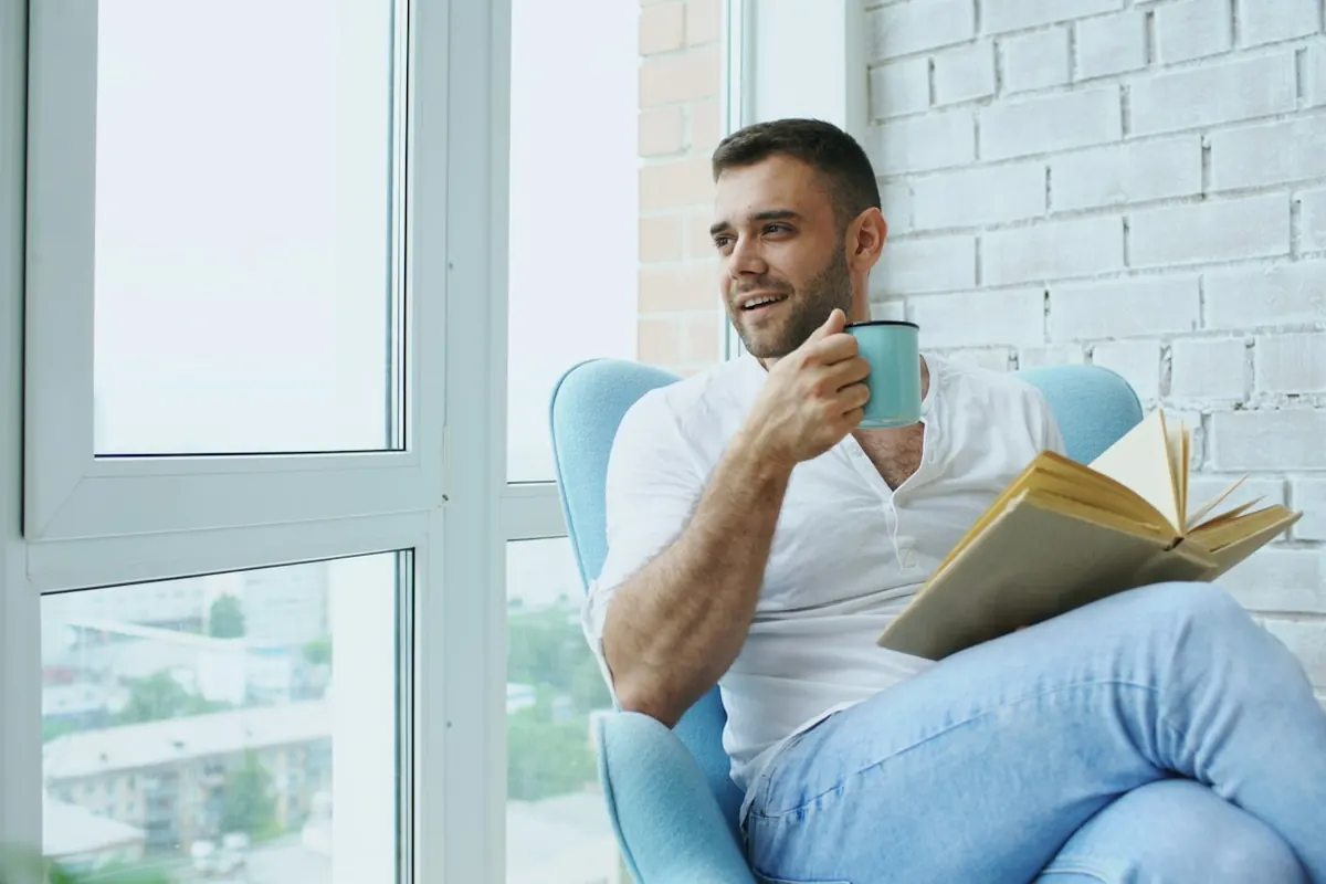 Man reading a book and drinking coffee by window